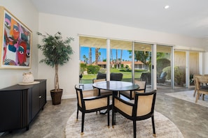 A dining area with a round table, four chairs, a potted plant, a cabinet, and a large window with a view of palm trees and a lawn. - Premier 3-Bedroom Condo in Coveted Indian Wells Country Club, Discounted Rates! (Indian Wells)