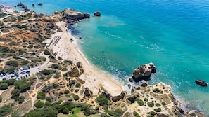An aerial view of a coastal area with a beach, rocky outcrops, and a parking lot with vehicles. - The Beach House - Villa walking to the beach (Albufeira)