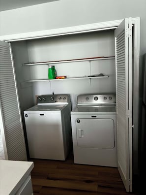 A laundry room with two washing machines and a dryer, a shelf with cleaning supplies, and a door leading to another room.