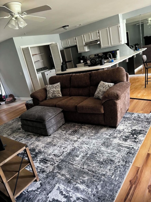 A living room with a brown sofa, patterned cushions, a grey and black rug, a wooden coffee table, and a ceiling fan.