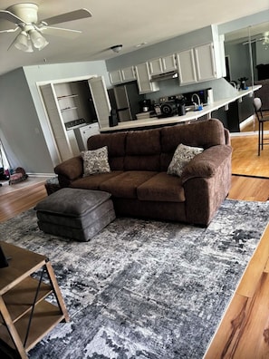 Un salon avec un canapé marron, des coussins à motifs, un tapis gris et noir, une table basse en bois et un ventilateur de plafond.