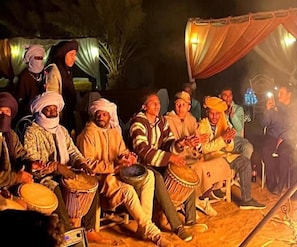 A group of people seated outdoors, some playing drums. - Moroccan Desert Camp (Rissani)