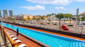 Une piscine avec une terrasse en bois et des chaises longues, une rue animée avec des voitures et un camion rouge, et de grands immeubles en arrière-plan.
