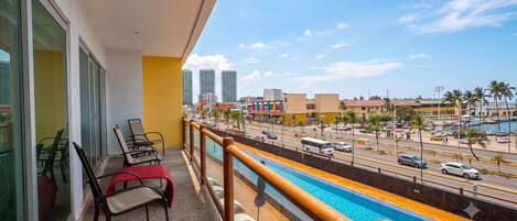 Un balcón con vista a una piscina, una calle con coches y edificios altos al fondo.