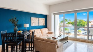Un salon moderne avec un mur d’accent bleu, un coin repas avec une table et des chaises, un canapé beige et une grande fenêtre donnant sur des palmiers et une marina.