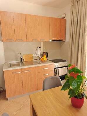 A compact kitchen with wooden cabinets, a sink, a stove, and a dining area with a table and a potted plant. - 2BR Near Beach  Big Terrace Free Parking (Golem)
