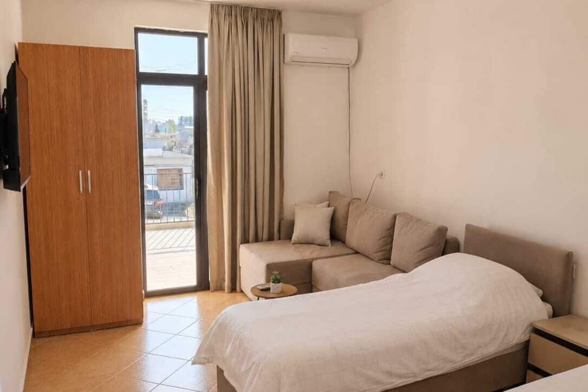 A hotel room with a bed, a sofa, a small table, a wardrobe, and a sliding glass door to the outside.