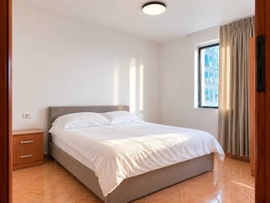A bedroom with a bed, a nightstand, a window with curtains, and a view of buildings outside. - 2BR Near Beach  Big Terrace Free Parking (Golem)