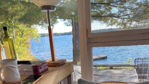 Une table en bois avec une lampe, des livres, une tasse et une bougie. Une chaise avec un coussin sur lequel est écrit « SAUTE DANS LE LAC » est placée devant une fenêtre donnant sur un lac.