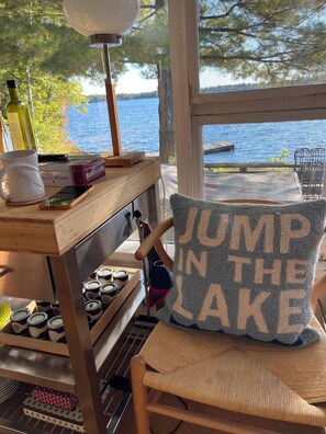 A wooden table with a lamp, books, a mug, and a candle. A chair with a cushion that says 'JUMP IN THE LAKE' is in front of a window with a view of a lake. - LAKEFRONT 3BDR CABIN WITH PRIVATE DOCKS & DECKS - MUSKOKA  NEAR PORT CARLING (Port Severn)