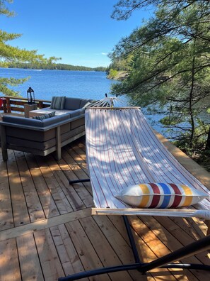 Un hamac sur une terrasse en bois donnant sur un lac, avec un canapé et une lanterne.