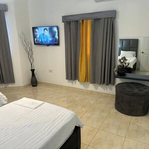 Quarto moderno com cama, televisão, janela com cortinas, um vaso e um pequeno banquinho.