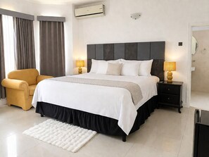 A modern hotel room with a large bed, two bedside tables with lamps, a sofa, a white rug, and an air conditioning unit. - Runaway Dreams Jamaica – Your Private Paradise Awaits (Runaway Bay)