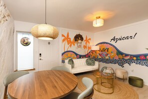 Une pièce avec une table ronde en bois, un canapé blanc, un luminaire suspendu en fibres tressées et une fresque colorée sur le mur.