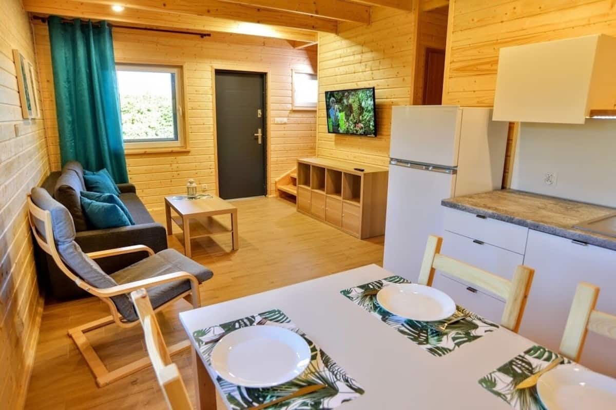 Una cabaña de madera con sofá, una mesa de comedor para dos, televisor y zona de cocina.