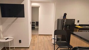 A compact kitchen with a refrigerator, a TV mounted on the wall, and a small dining area with a chair. - Sevillavenue (Ciudad de México)