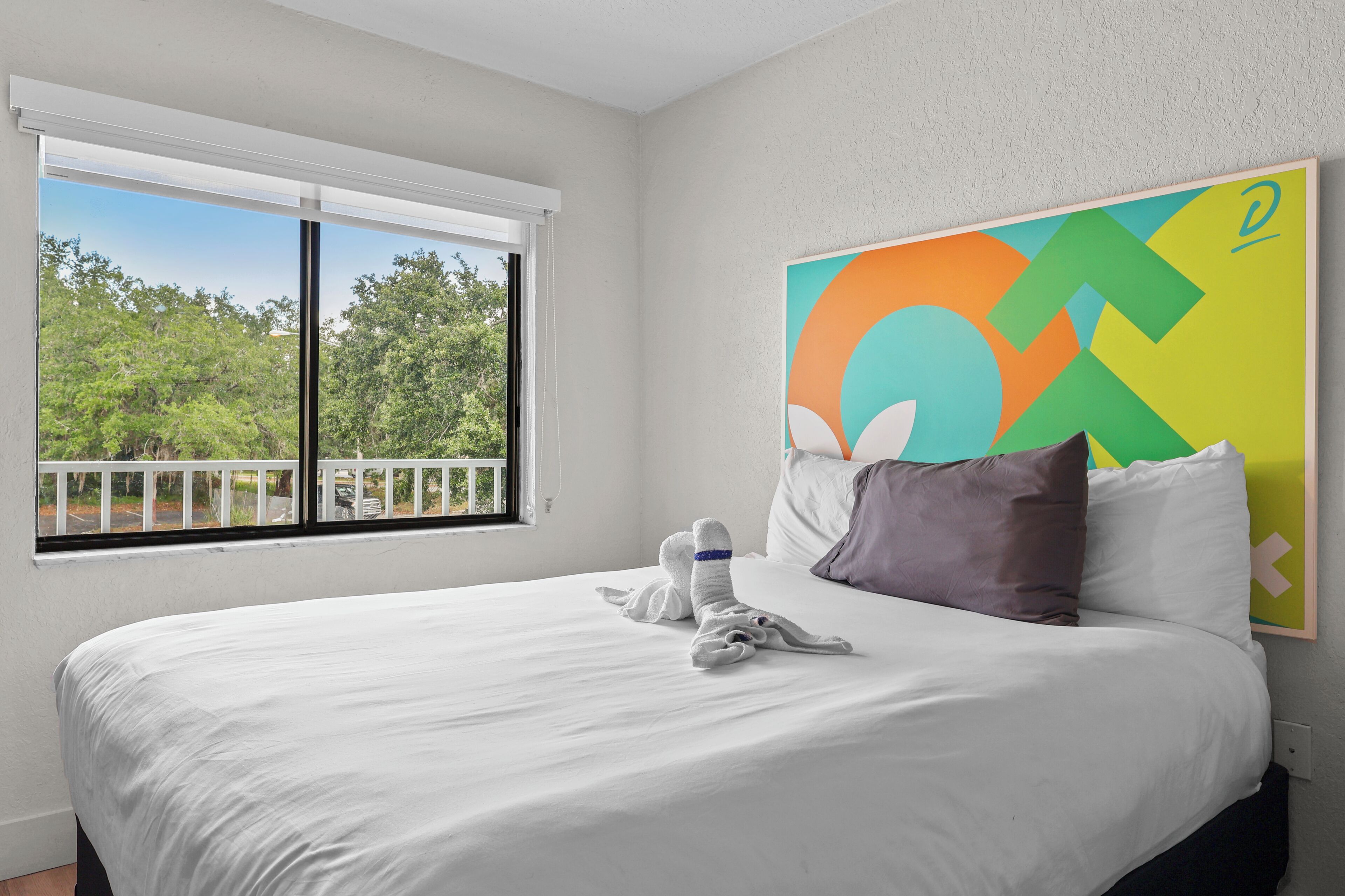 Une chambre d’hôtel avec un grand lit, une fenêtre donnant sur des arbres et une peinture abstraite aux couleurs vives accrochée au mur.