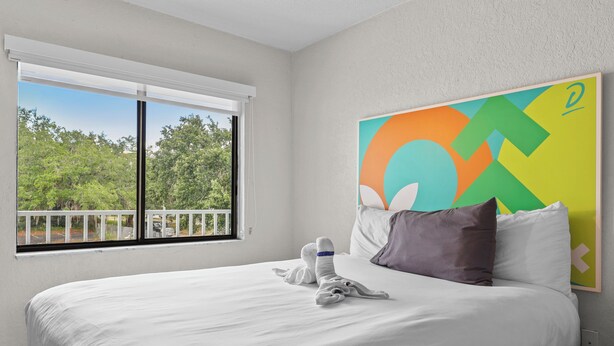 A hotel room with a large bed, a window with a view of trees, and a colorful abstract painting on the wall. - Budget Disney Room Pool & Parking (Kissimmee)