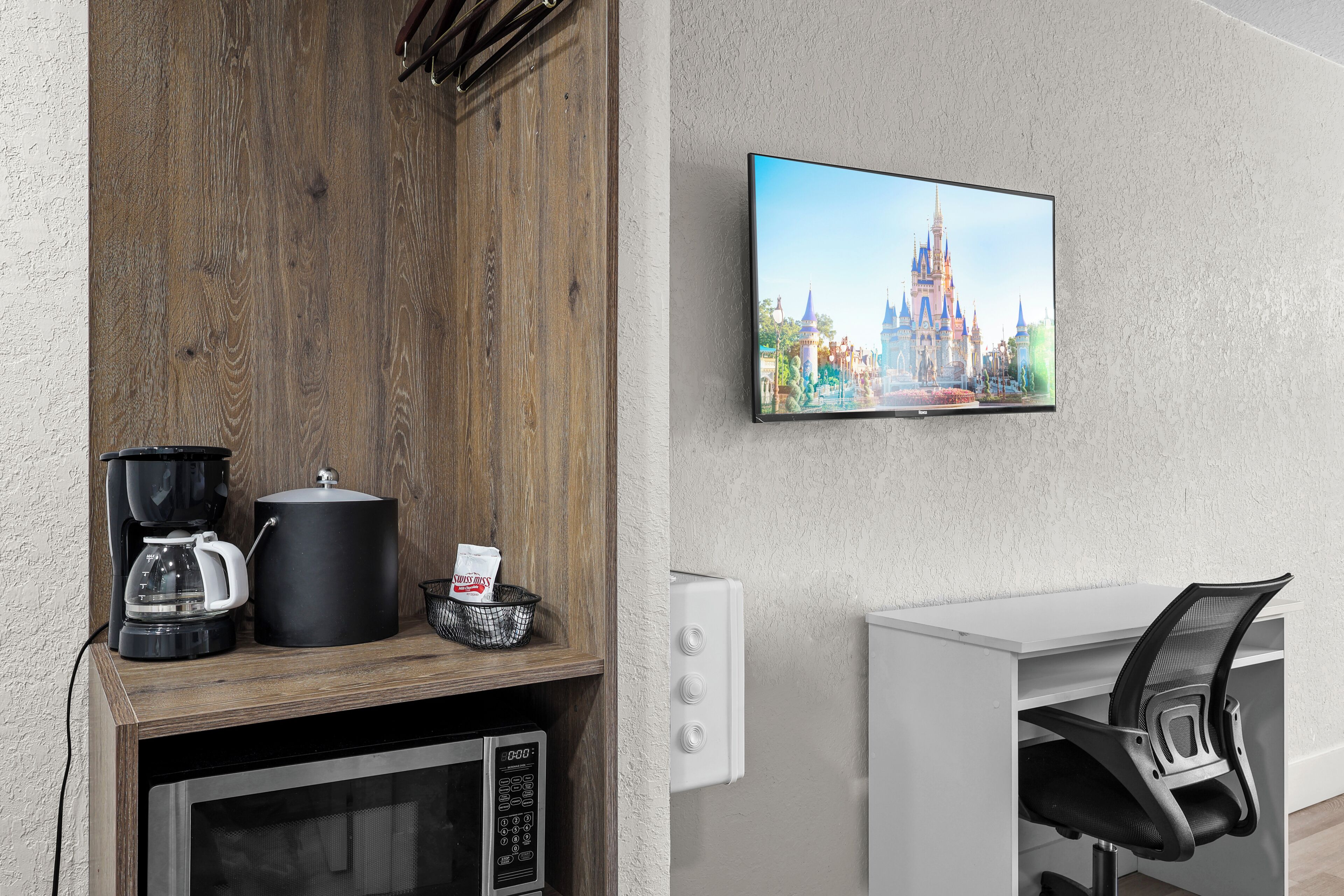 Une chambre moderne avec un meuble en bois, une machine à café, un four à micro-ondes, un téléviseur affichant une image de château et un bureau blanc avec une chaise.