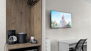 A modern room with a wooden cabinet, a coffee maker, a microwave, a TV displaying a castle, and a white desk with a chair. - Budget Disney Room Pool & Parking (Kissimmee)