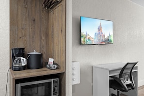 Una habitación moderna con un mueble de madera, una cafetera, un microondas, un televisor mostrando un castillo y un escritorio blanco con una silla.