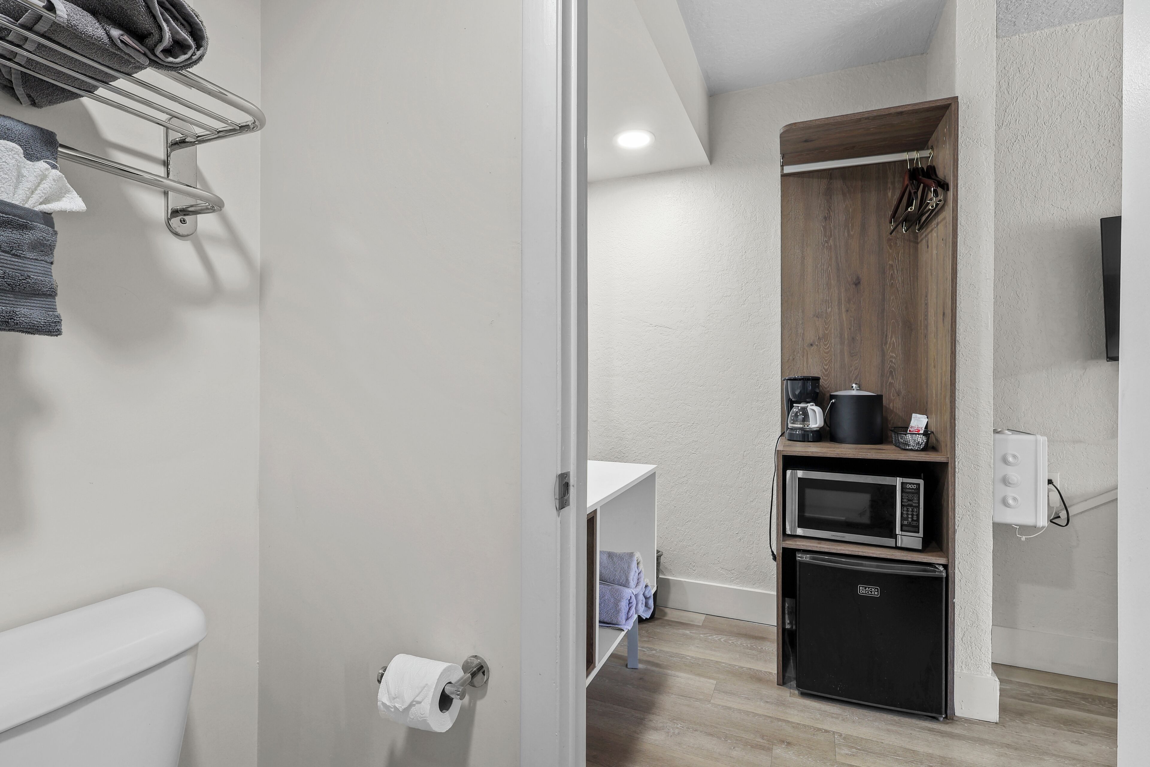 A compact bathroom with a toilet, a towel rack, a shelf with towels, a microwave, and a refrigerator.