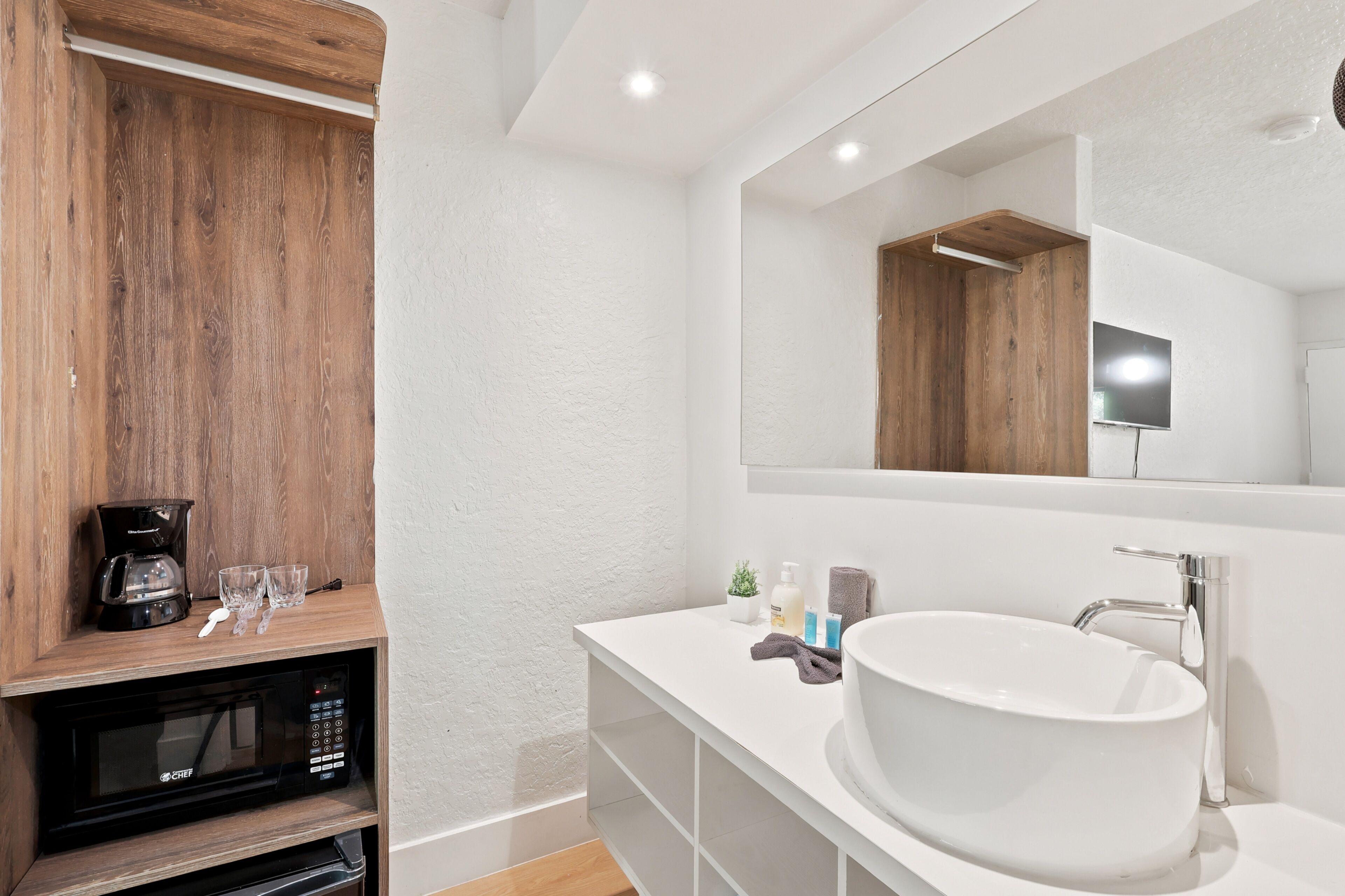 Une salle de bain moderne avec un lavabo blanc, un grand miroir, une étagère en bois sur laquelle se trouve une machine à café et un four à micro-ondes.