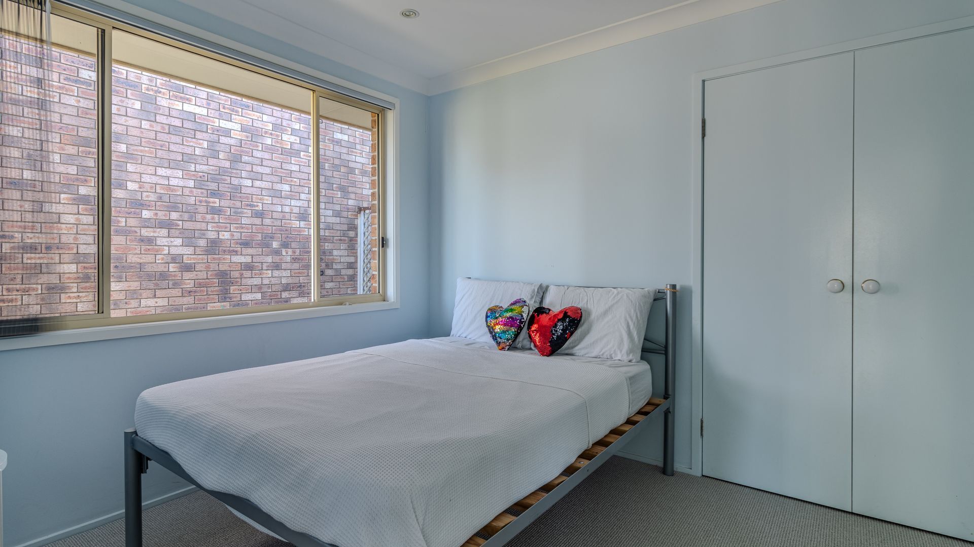 A bedroom with a single bed, a window with a brick wall view, and a closed door.