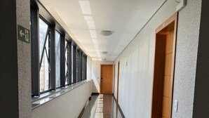 Un pasillo largo y estrecho con suelo pulido, ventanas amplias en un lado y puertas de madera en el otro.