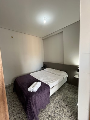 Een hotelkamer met een bed, een paars deken, een witte handdoek, een nachtkastje, een lamp en een plafondlamp.