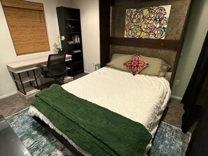 Ein Schlafzimmer mit Bett, Schreibtisch, Sessel, Bücherregal und einem Bild an der Wand.