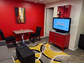 Ein Wohnzimmer mit roter Wand, einem Gemälde, einem Fernseher, der einen Sale anzeigt, einer roten Kommode und einem kleinen Tisch mit Stühlen.