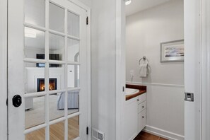 Una habitación con chimenea, suelo de madera, puerta blanca, baño con lavamanos y toallero, y un cuadro enmarcado en la pared.