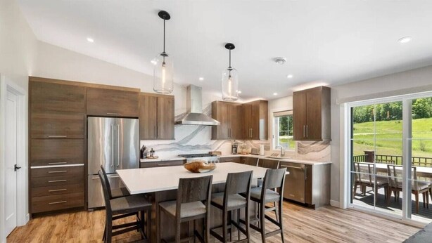 A modern kitchen with a central island, wooden cabinets, stainless steel appliances, and a view of a green landscape through large sliding glass doors.