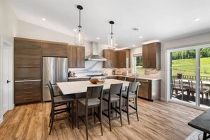 A modern kitchen with a central island, wooden cabinets, stainless steel appliances, and a view of a green landscape through large sliding glass doors.