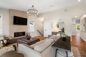 A modern living room with a fireplace, a large flat-screen TV, a dining area with a table and chairs, and a staircase leading to another level.
