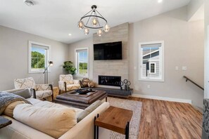 A spacious living room with a fireplace, a large flat-screen TV mounted on the wall, and a modern chandelier.