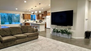 A modern living room with a sofa, a flat-screen TV mounted on the wall, and a kitchen in the background. - HandyFarms.com: California Organic Farm Stay on a Red Walnut Orchard Nut Store (Corning)
