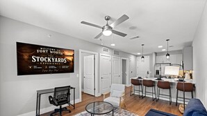 A modern living room with a sofa, a glass coffee table, a desk with a chair, a kitchen area with bar stools, and a ceiling fan. - New Modern King bed+Pool+Gym+Parking+Stockyards (Fort Worth)