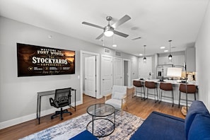 Un salon moderne comprenant un canapé, une table basse en verre, un bureau avec une chaise, un coin cuisine avec des tabourets de bar et un ventilateur de plafond.