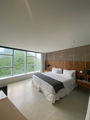 Une chambre d’hôtel moderne avec un grand lit, une tête de lit en bois, une table de chevet, un bureau et une vue sur un paysage montagneux par de grandes fenêtres.