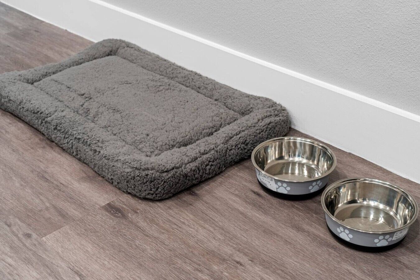 A gray pet bed with two metal bowls beside it.