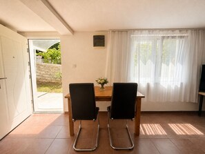 Ein Zimmer mit einer Tür zu einem Außenbereich, einem Tisch mit zwei Stühlen, einer kleinen Vase mit Blumen und einem Fernseher auf einem Schrank.