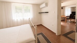 Une chambre à coucher comprenant un lit, une fenêtre avec des rideaux, un appareil de climatisation, un bureau avec une chaise et une porte donnant sur une autre pièce.