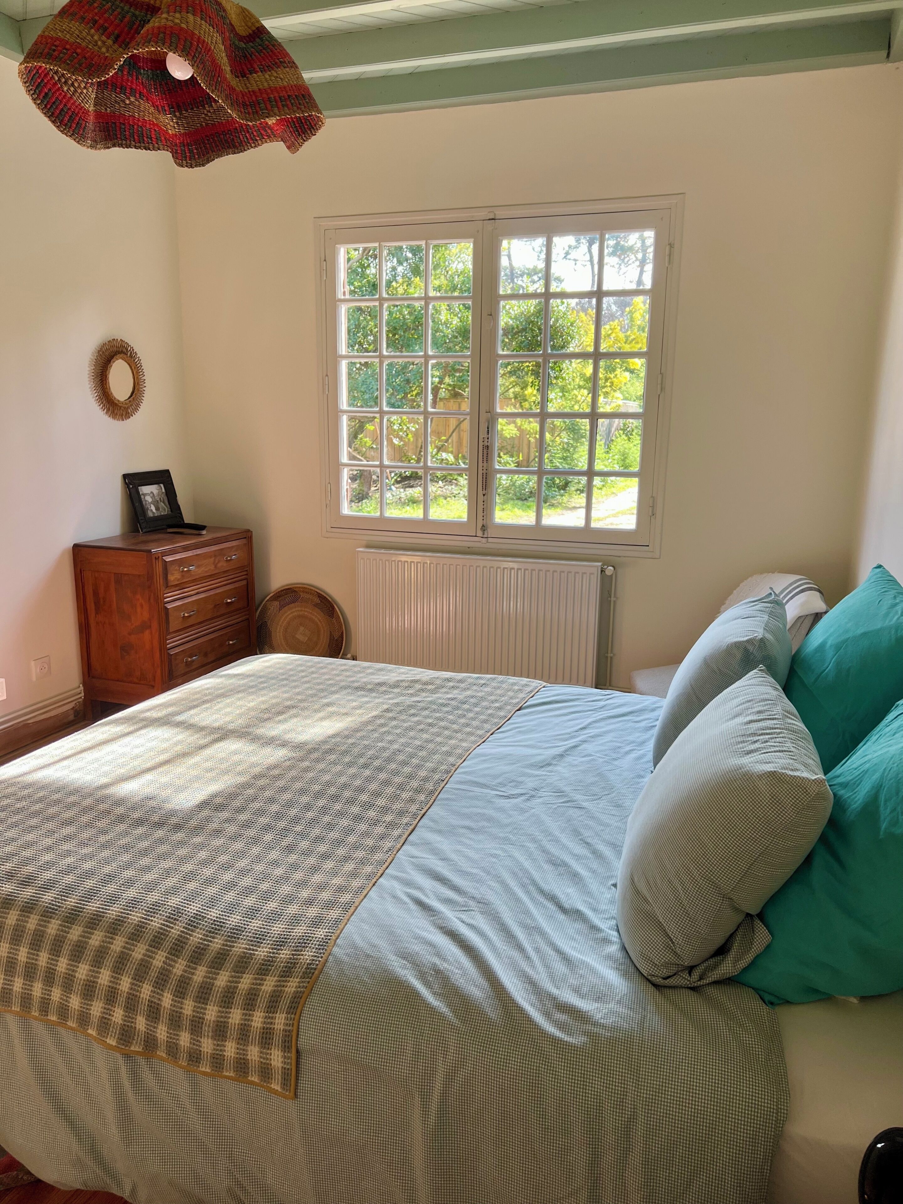 A bedroom with a bed, a window, a radiator, a wooden dresser, and a wall-mounted mirror.