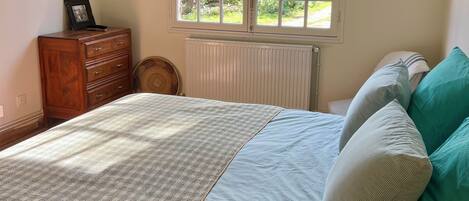A bedroom with a bed, a window, a radiator, a wooden dresser, and a wall-mounted mirror.