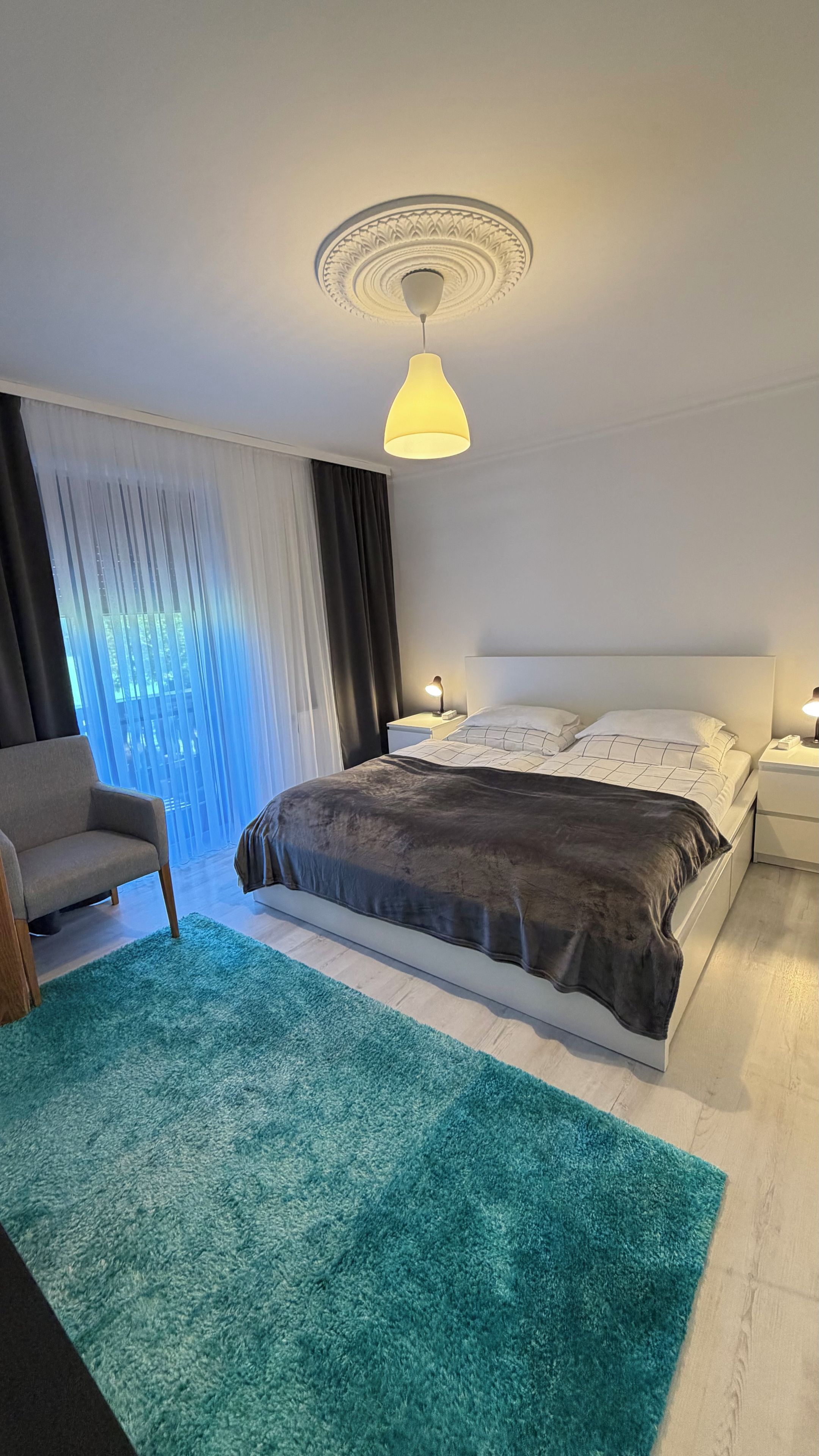 A hotel room with a bed, bedside tables, a chair, a ceiling light fixture, and a window with curtains.