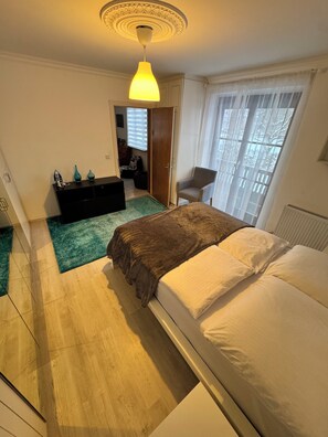 Une chambre à coucher avec un lit, une commode, une chaise, un radiateur et une fenêtre avec des rideaux.