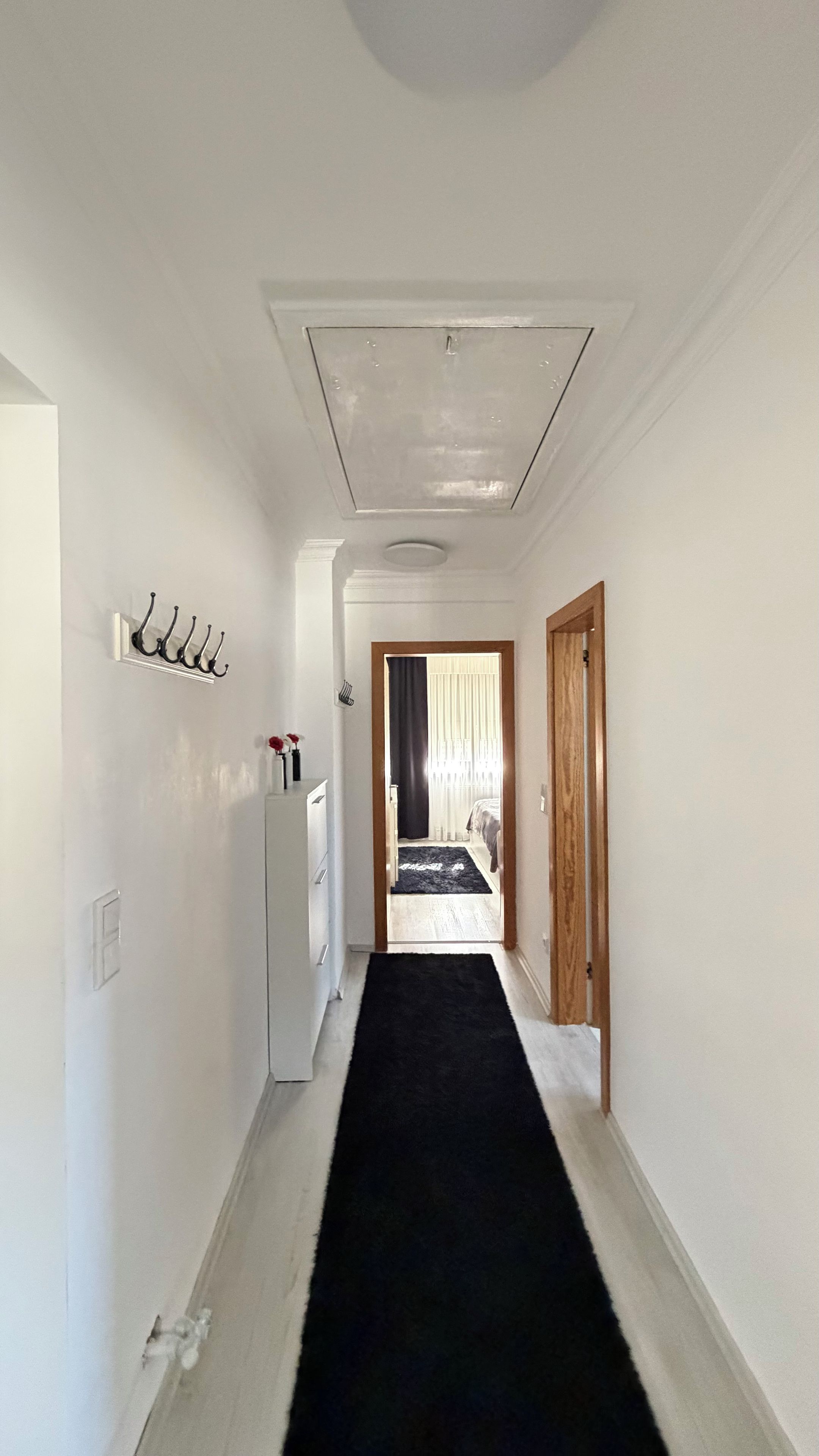 A narrow hallway with a black carpet leading to a room with a bed and a window with curtains.