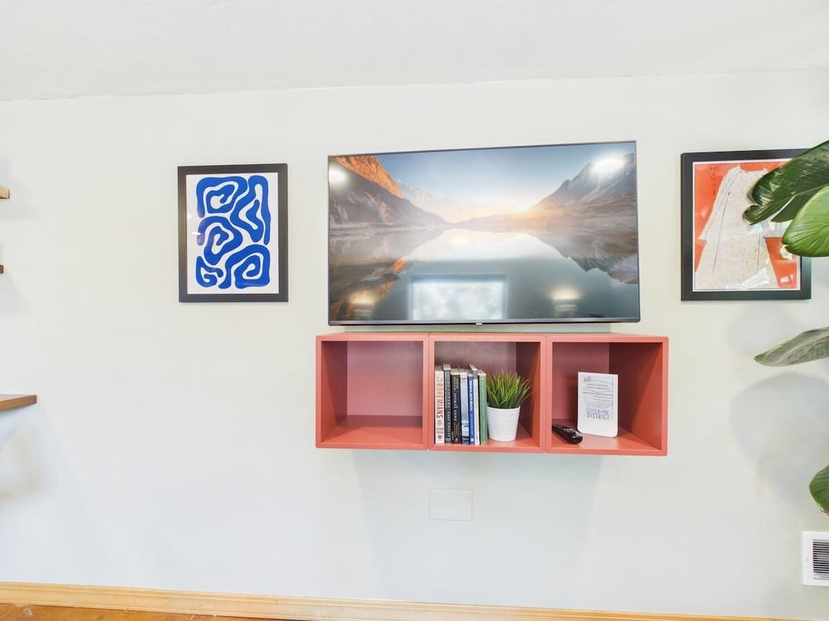 Ein Flachbildfernseher an einer weißen Wand, begleitet von zwei gerahmten Kunstwerken und einem rosa Regal mit Büchern und einer Topfpflanze.
